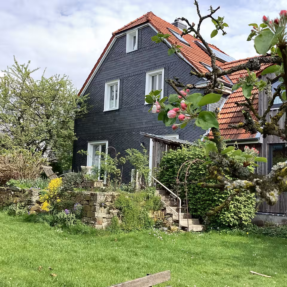 Hinter einem Garten mit einer Steinmauer und einem blühenden Apfelbaum im Vordergrund steht ein Haus mit Schieferverkleidung und rotem Dach.