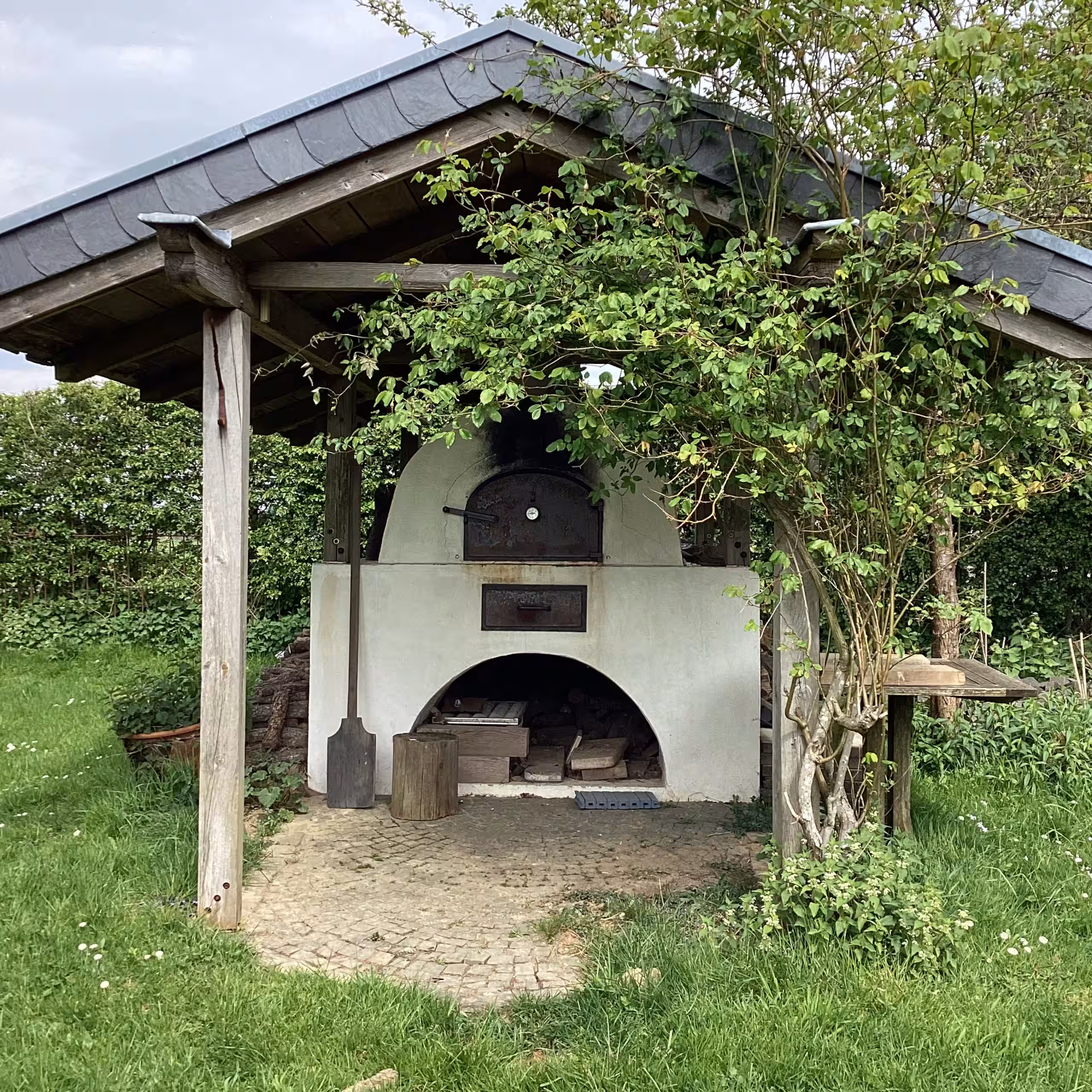 Ein weißer Holzofen für Pizza im Freien steht unter einem Holzunterstand in einem grasbewachsenen Garten.