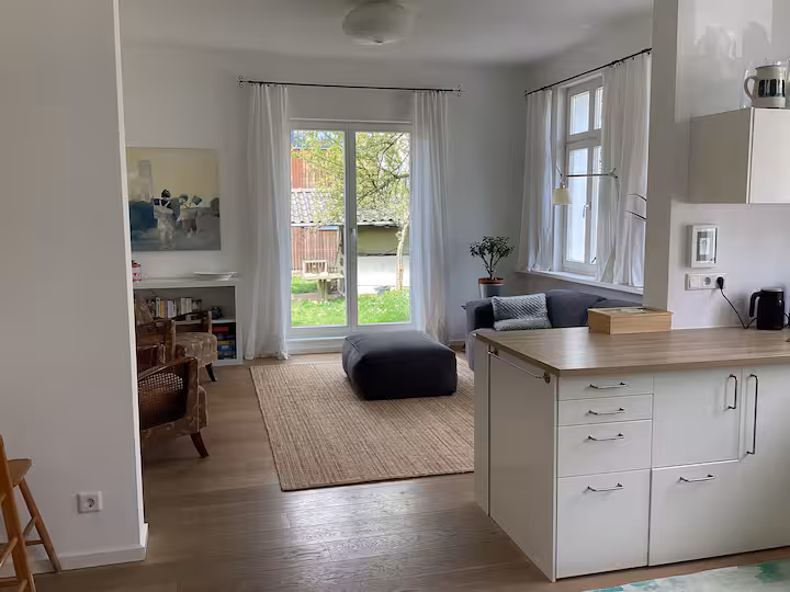 Wohnzimmer mit einer Kücheninsel im Vordergrund, die zu einer Sitzecke mit Stühlen, einem Sofa und einer Glastür führt, die zum Garten hin öffnet.
